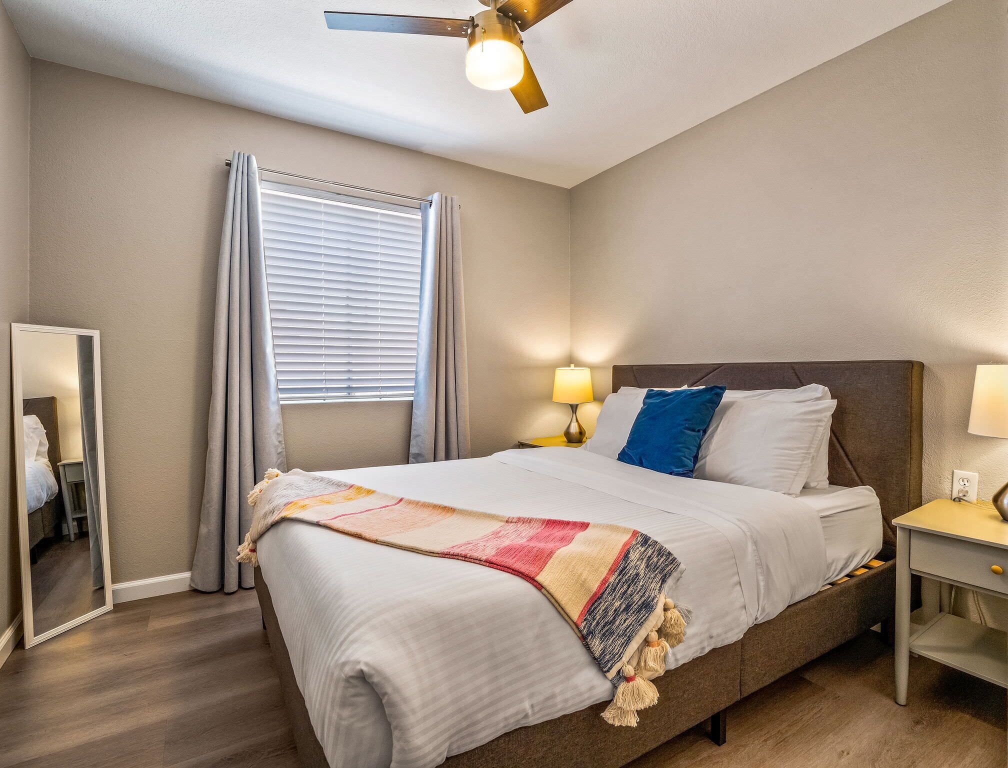 A hotel room with a bed, a ceiling fan, a window with curtains, a bedside table with a lamp, and a mirror.