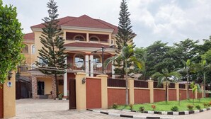A two-story house with a red-tiled roof, a gated entrance, and a well-maintained garden with various plants and flowers. - Royale Crown Villa Apartment (Abuja)