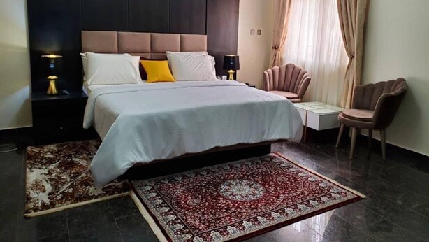 A hotel room with a large bed, a headboard, a nightstand with a lamp, a chair, a small table, and a patterned rug. - Royale Crown Villa Apartment (Abuja)