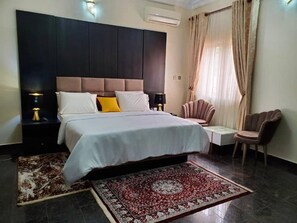 A hotel room with a large bed, a headboard, a nightstand with a lamp, a chair, a small table, and a patterned rug.