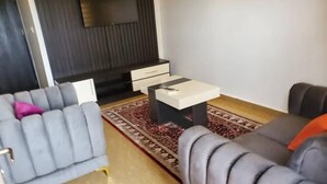 A modern living room with a grey sofa, a smaller grey armchair, a coffee table, and a TV mounted on a dark wall. - Royale Crown Villa Apartment (Abuja)