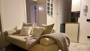 En beige soffa med kuddar och en pläd, en liten pall, en vit dörr med ett fönster och ett vitt kylskåp.