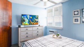 Ein Schlafzimmer mit einem Bett, einem Fernseher auf einer Kommode, einem Deckenventilator und einem Fenster mit Jalousien.