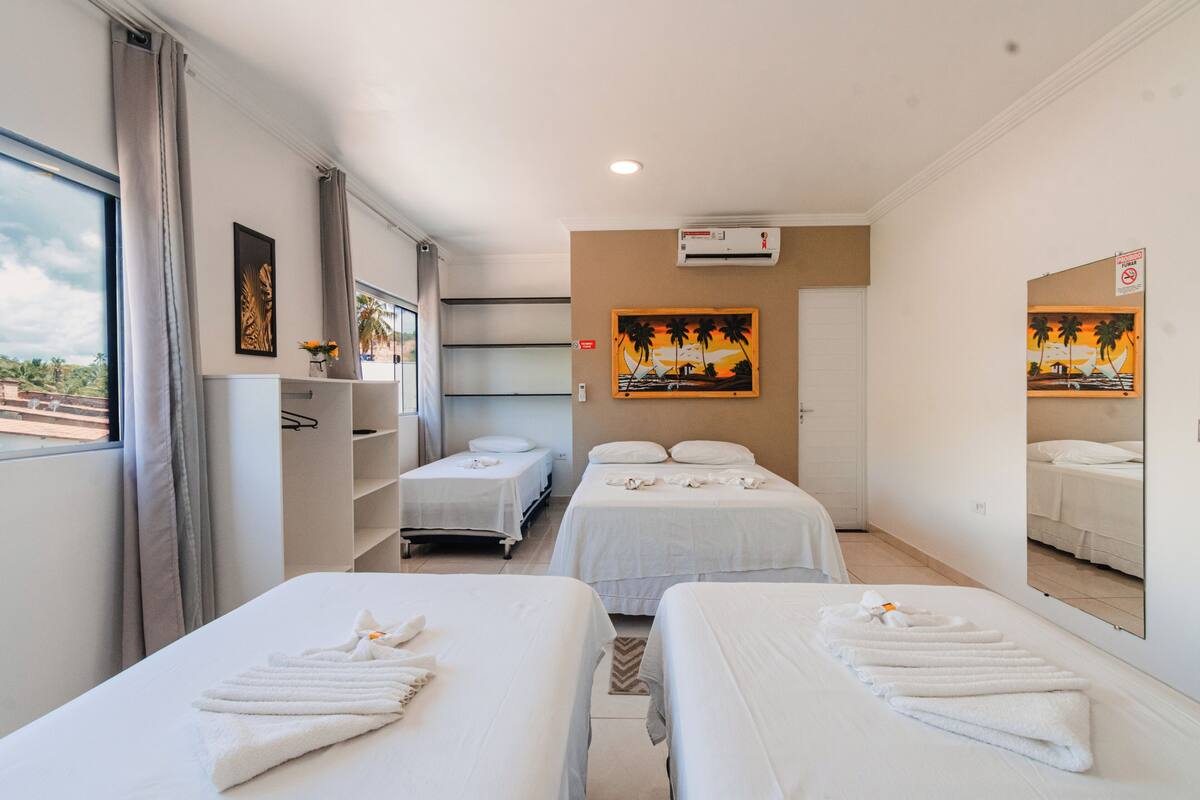 Quarto de hotel com duas camas, uma janela grande com cortinas, um quadro na parede e um aparelho de ar condicionado.
