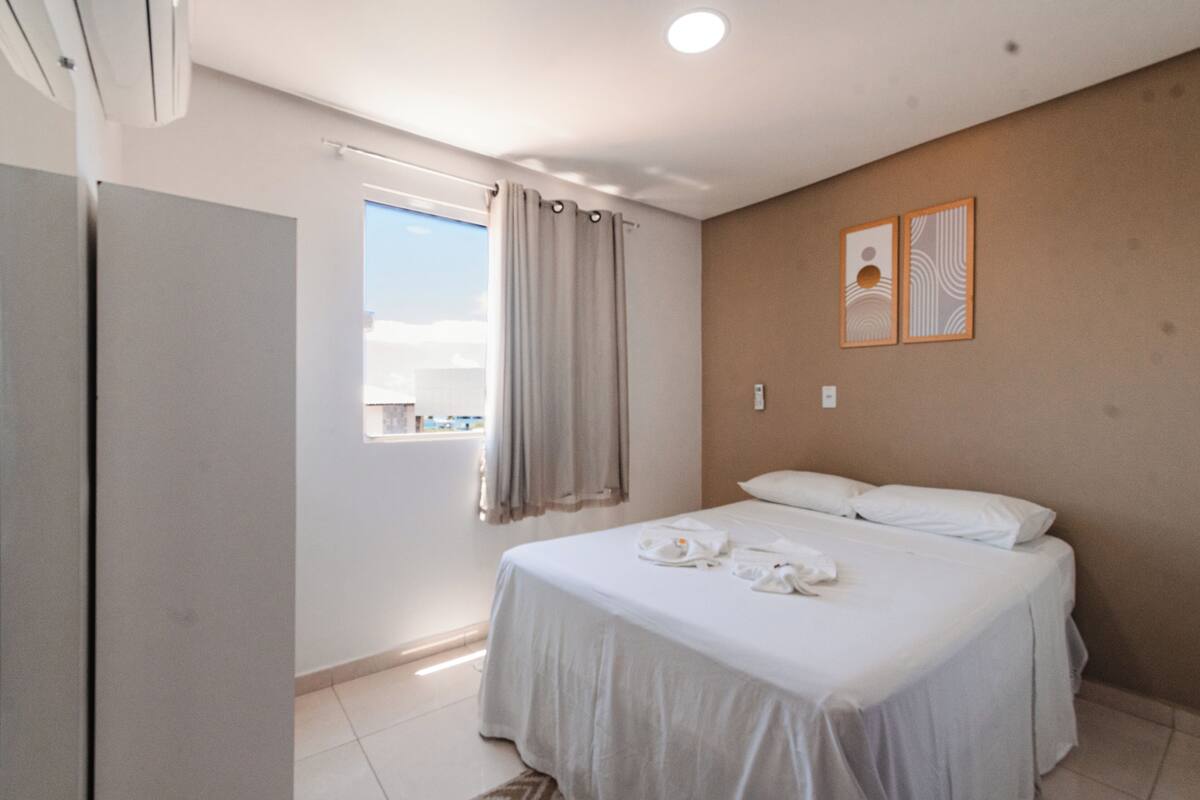 Quarto de hotel com cama, janela com cortinas, obra de arte na parede e iluminação no teto.