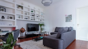 A modern living room with a grey sofa, a flat-screen TV, and white shelving with various decorative items. - Italianway - Cristoforo Colombo 5B (Milan)