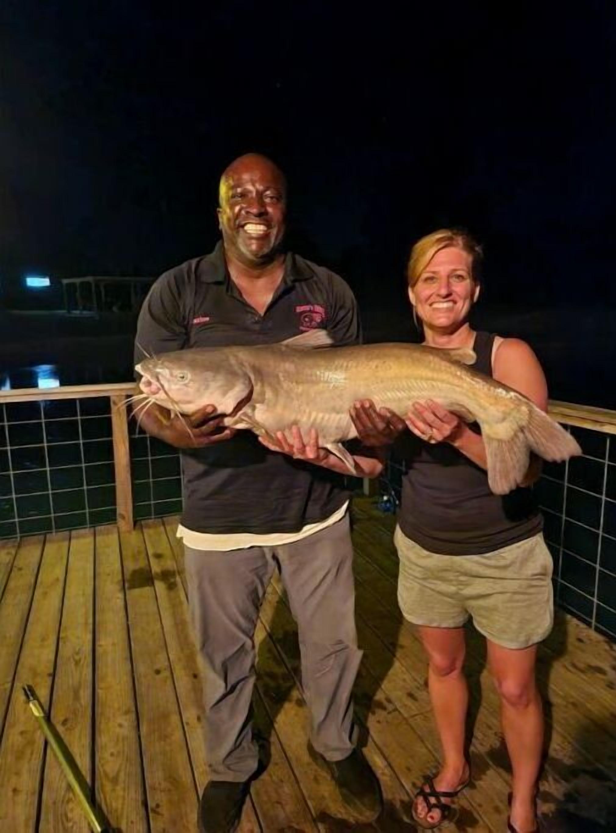 Two people holding a large fish on a wooden deck at night.