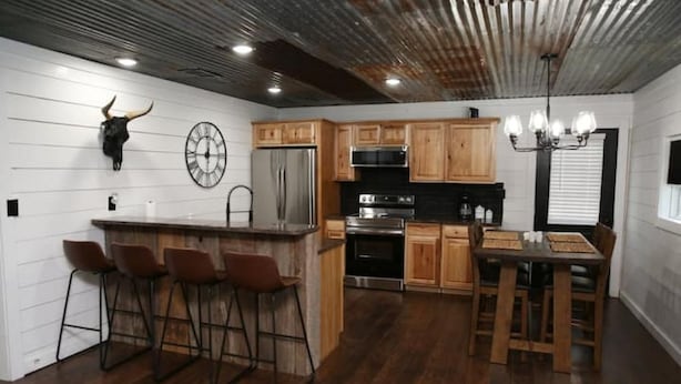 A kitchen with wooden cabinets, a stainless steel refrigerator, a clock, and a bar area with stools. - Hidden Gem Cottages (Palestine)