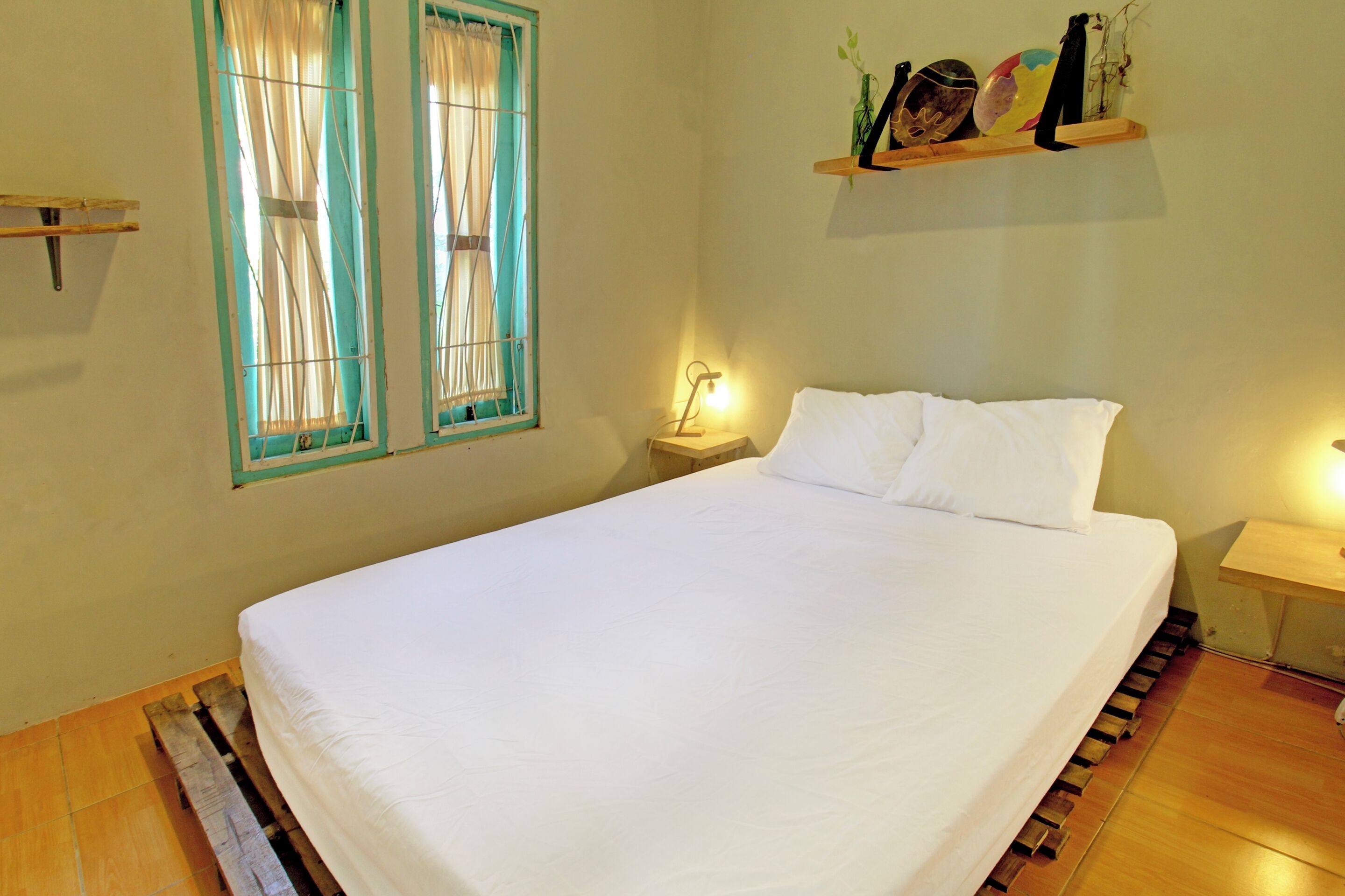 A neatly made bed in a room with wooden flooring, two windows with curtains, a bedside table with a lamp, and a shelf with decorative items.