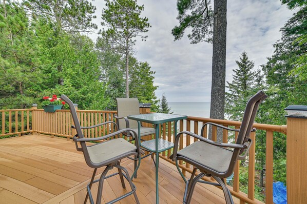 A wooden deck with outdoor furniture, surrounded by trees and overlooking a body of water.