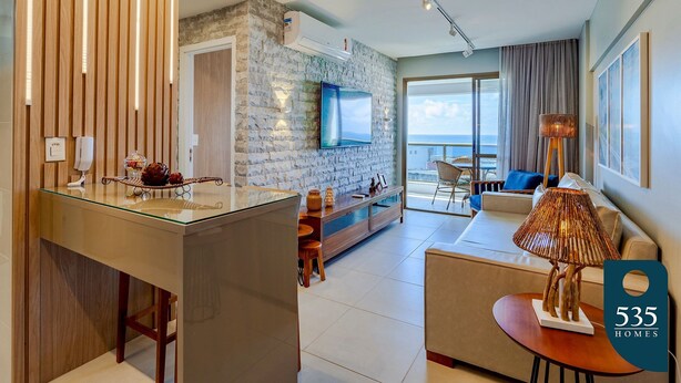 Een moderne woonkamer met een salontafel met glazen blad, een bank en een televisie bevestigd aan een stenen muur. Er is een balkon met uitzicht op zee.