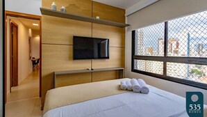 A modern hotel room with a bed, a TV mounted on the wall, a window with a city view, and a small table with towels. - Beautiful and cozy with a panoramic view in Barra (Salvador)