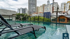 A rooftop swimming pool with lounge chairs, surrounded by urban buildings and greenery. - Beautiful and cozy with a panoramic view in Barra (Salvador)