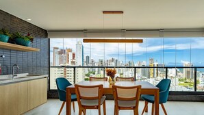 A modern dining area with a city view, featuring a wooden table and chairs, a vase with flowers, and a large window overlooking the cityscape. - Beautiful and cozy with a panoramic view in Barra (Salvador)