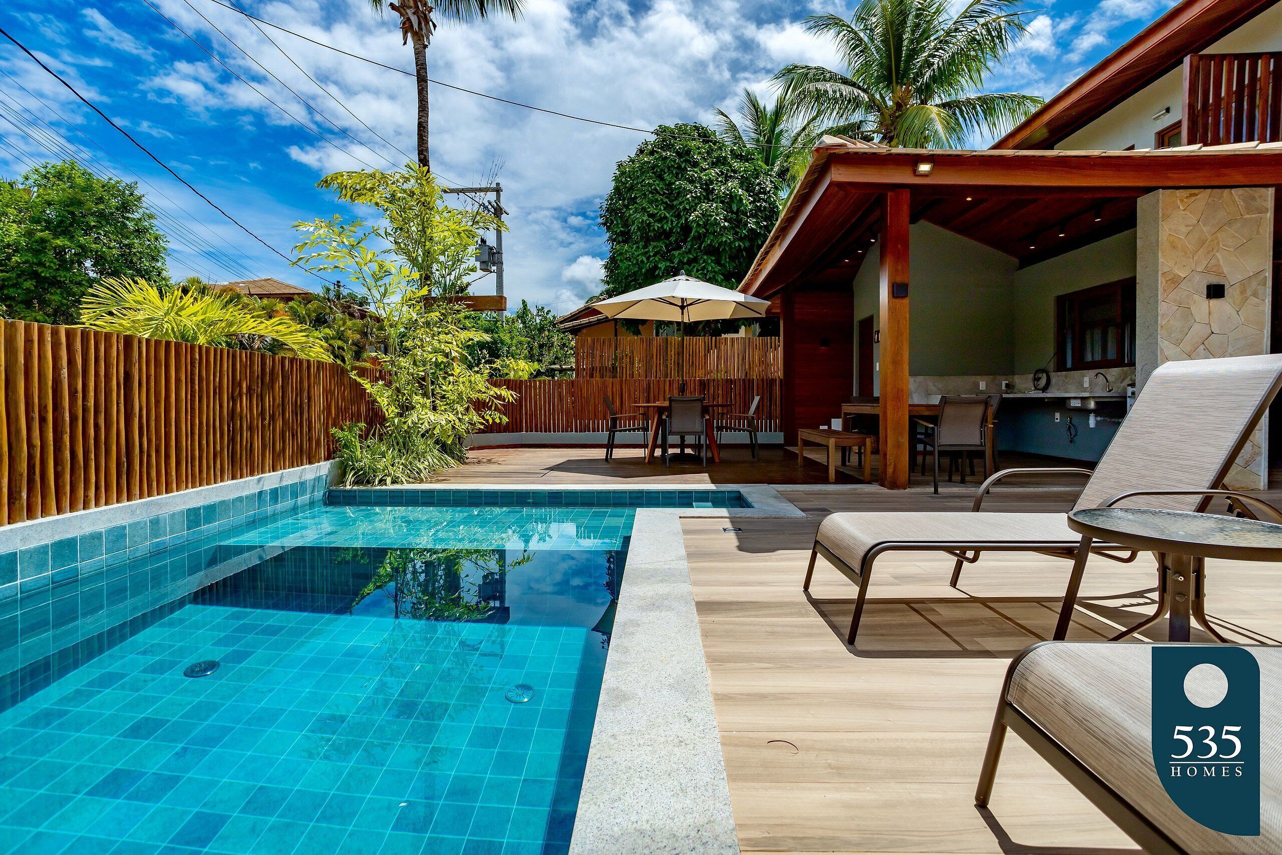 A swimming pool with lounge chairs, a wooden deck, and a covered outdoor area with a table and chairs.