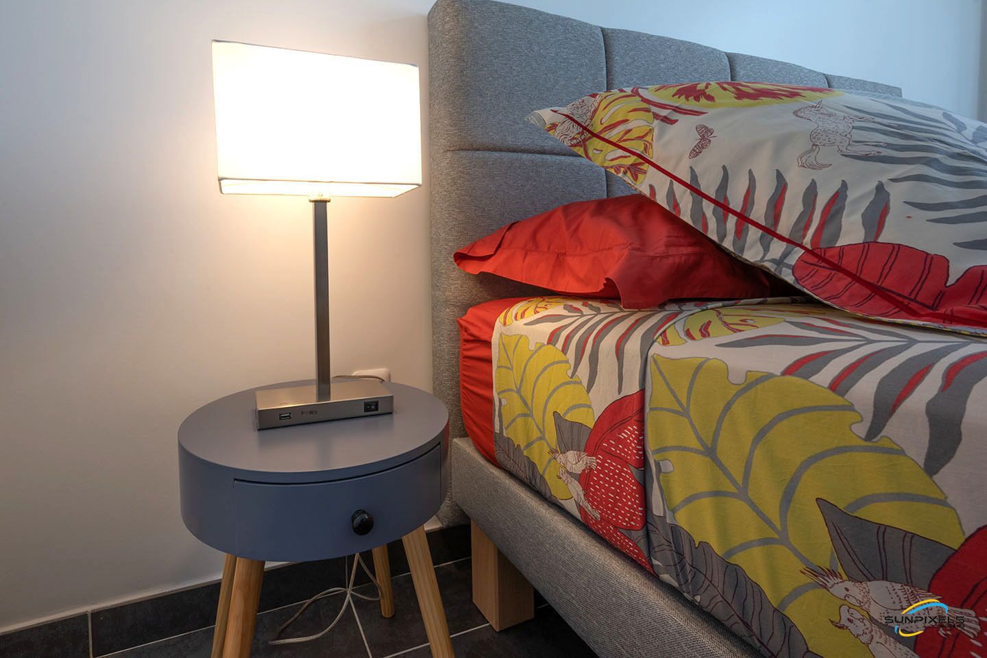 Un lit bien fait, avec une literie aux couleurs vives, une lampe de chevet et une petite table sur laquelle se trouve un appareil.