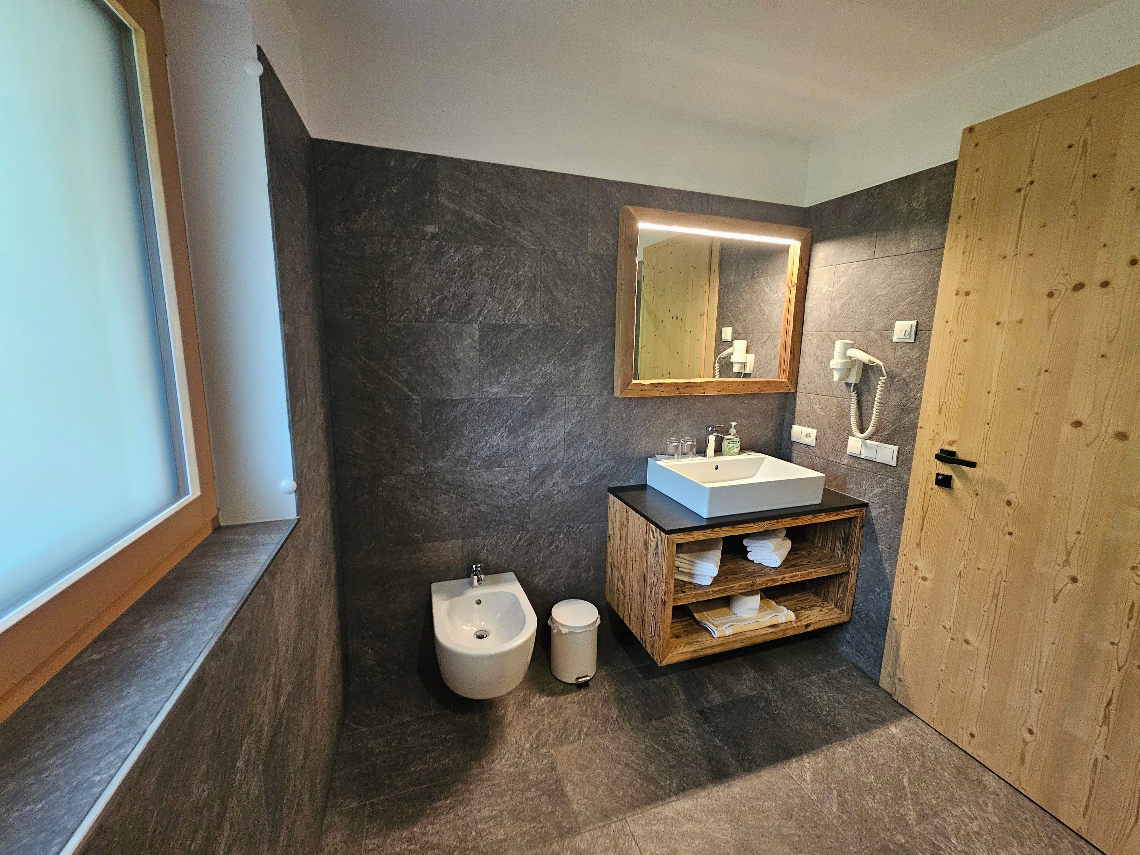 A modern bathroom with a dark stone wall, a wooden vanity with a sink, a bidet, and a mirror with integrated lighting.