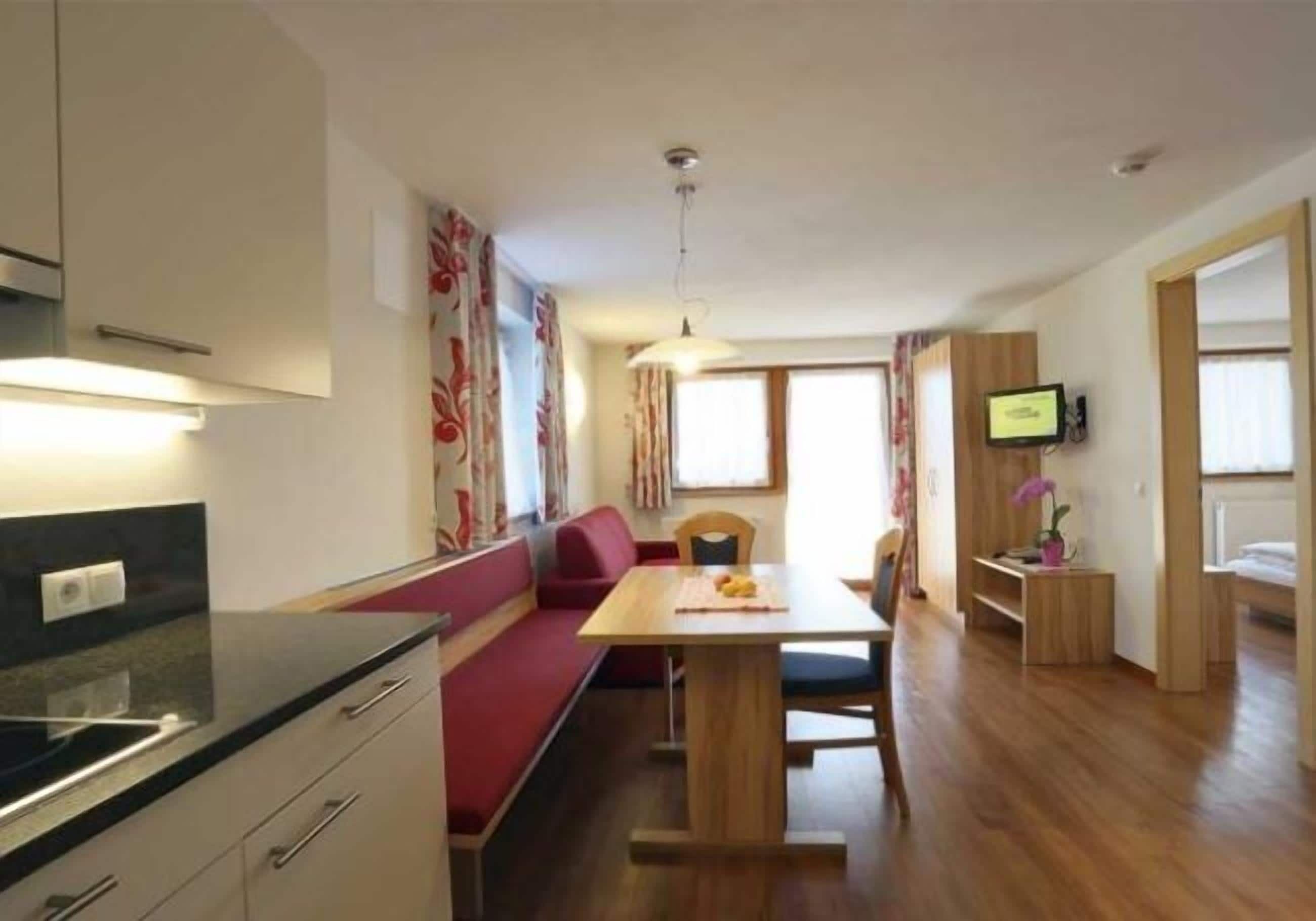 A modern kitchen with a built-in oven, a dining area with a wooden table and red seating, and a TV on the wall.
