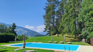 Ein Schwimmbecken mit Liegestühlen und Sonnenschirmen, umgeben von Grünflächen und Bergen.