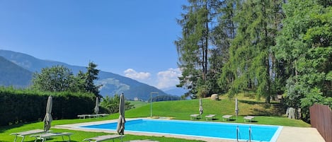 Ein Schwimmbecken mit Liegestühlen und Sonnenschirmen, umgeben von Grünflächen und Bergen.