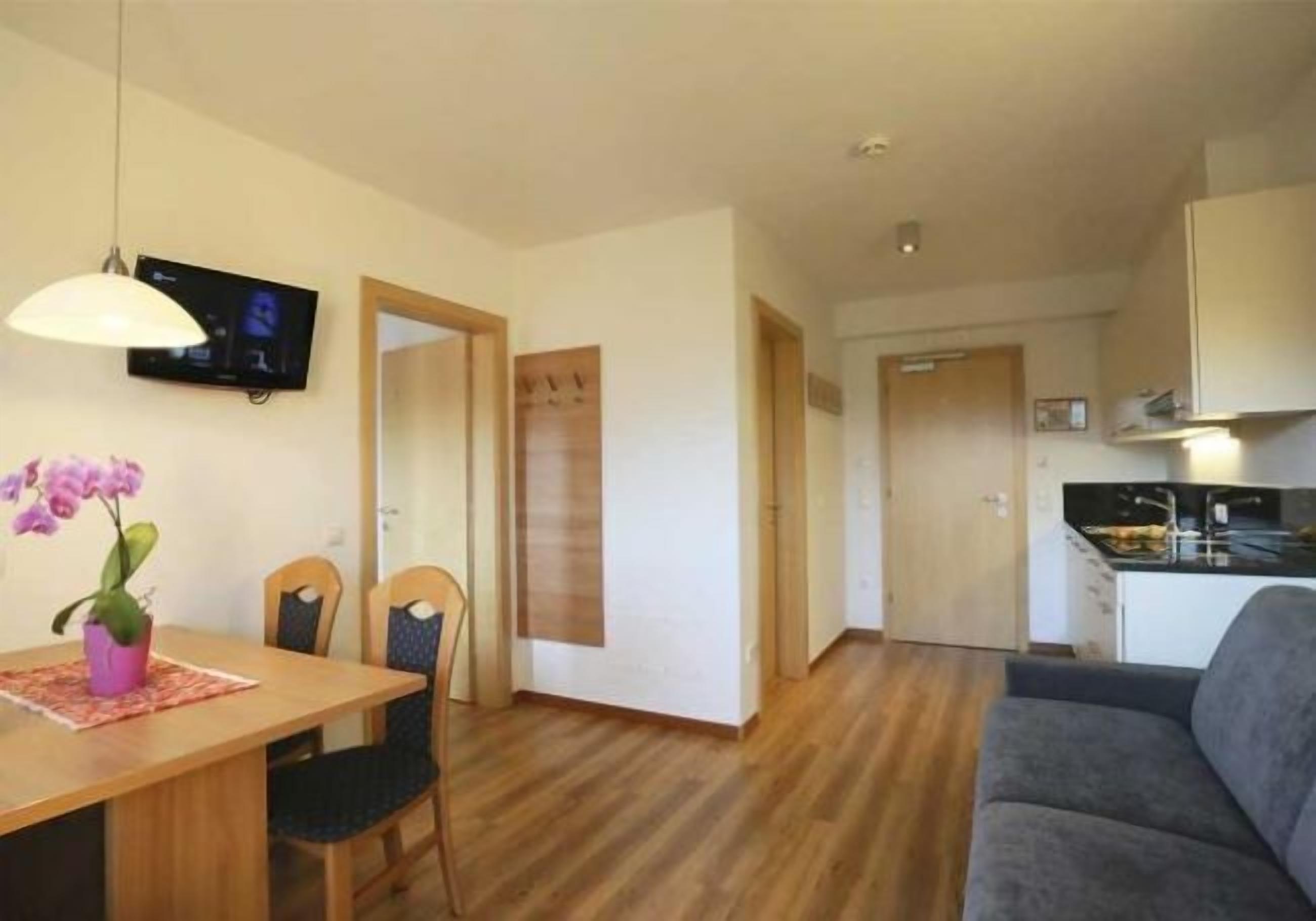 Un espacio habitable compacto con comedor, un sofá gris, una cocina y un televisor montado en la pared.