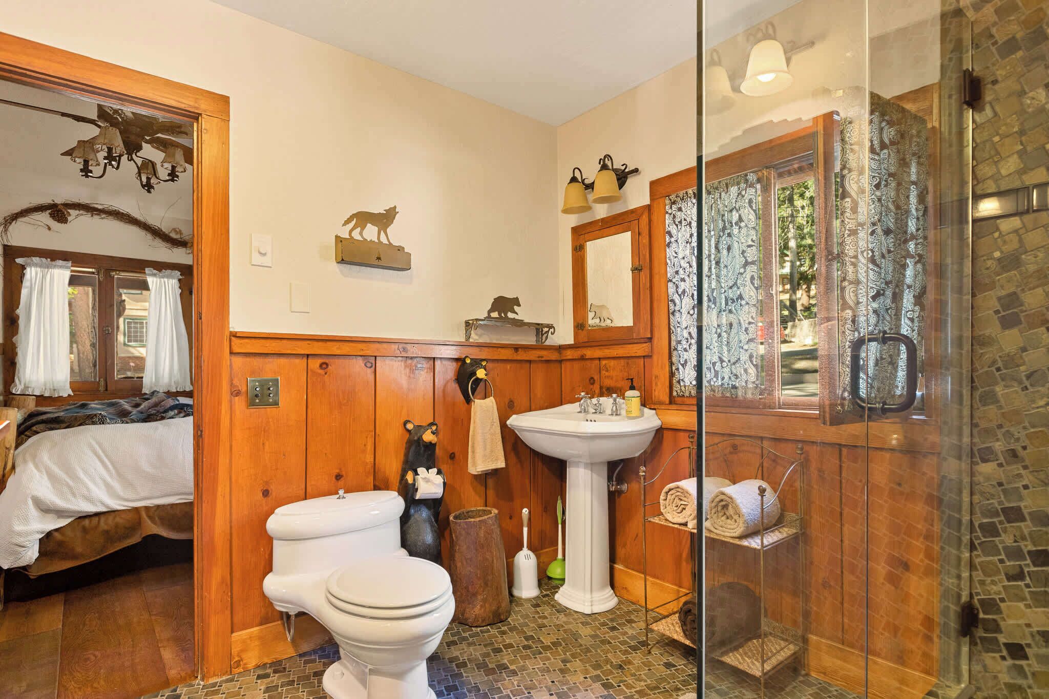 A bathroom with a wooden vanity, a white sink, a glass shower enclosure, and a bed in the adjacent room.