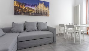 Uma sala de estar moderna com um sofá cinzento, cadeiras brancas e uma mesa de jantar. Um quadro grande de um edifício histórico está na parede.
