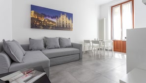 A modern living room with a grey sofa, a dining table with chairs, and a large window with curtains.
