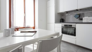 Eine moderne Küche mit weißen Schränken, einem Esstisch und Stühlen. Ein Fenster mit Vorhängen, ein Wasserkocher und eine Kaffeemaschine stehen auf der Arbeitsplatte.