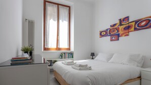 Ein Schlafzimmer mit einem Bett, einem Fenster mit Vorhängen, einem Nachttisch mit Büchern, einer Lampe und einer Wand mit farbenfroher Kunst.