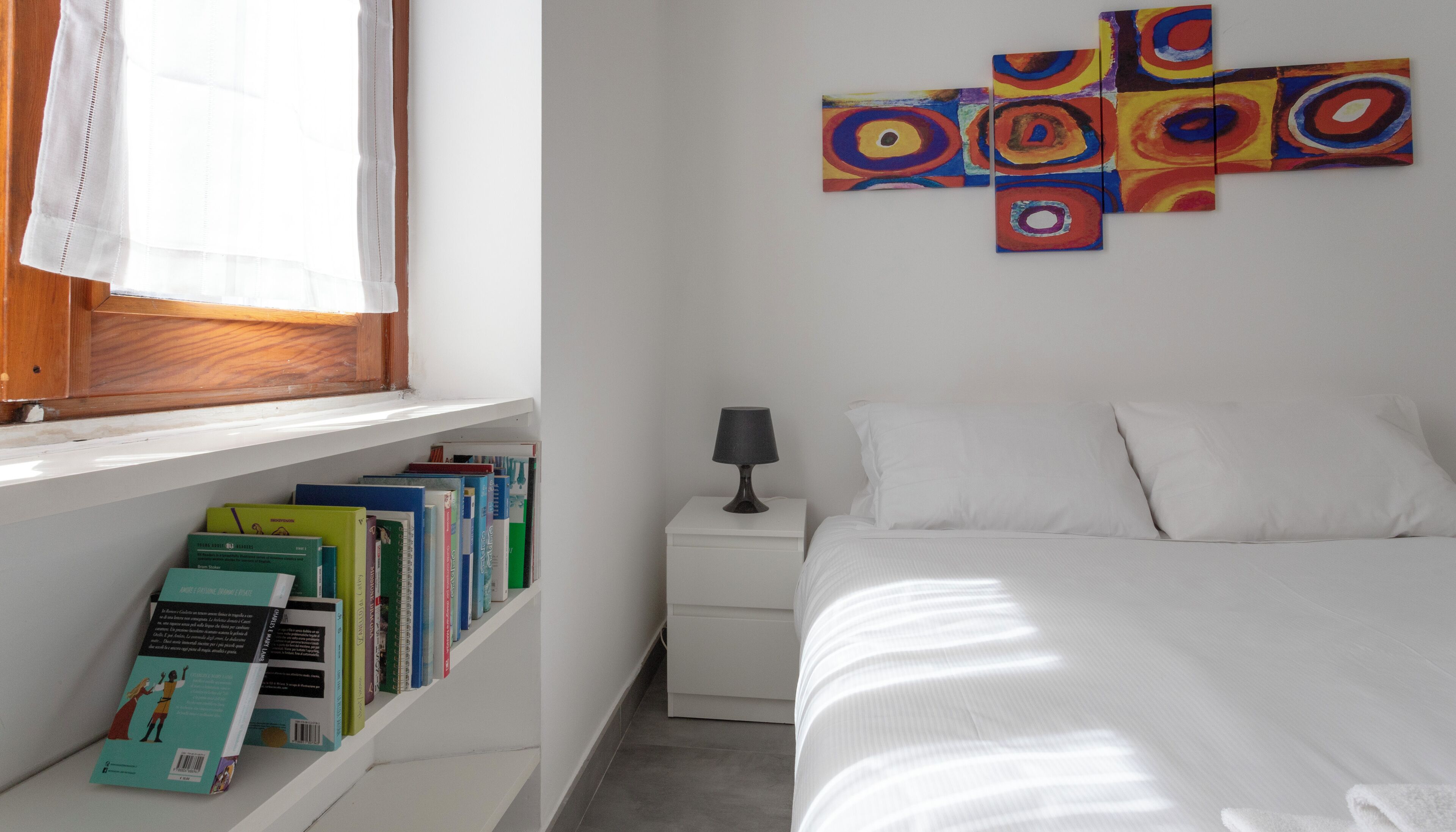 Une chambre à coucher comprenant un lit, une table de chevet, une lampe, une fenêtre avec des rideaux et une bibliothèque remplie de livres.