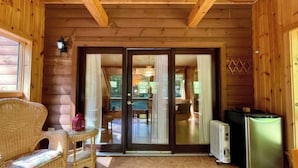 A wooden cabin interior with a wicker chair, a small table, a refrigerator, and a sliding glass door leading to a dining area. - Log Cabin w/  Full Garage, FirePit, BBQ, cozy gas heat Stove (Pocono Lake)
