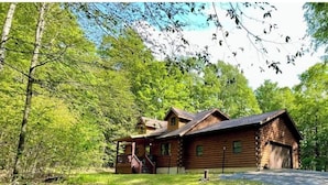A rustic log cabin with a porch and a garage, surrounded by dense green trees. - Log Cabin w/  Full Garage, FirePit, BBQ, cozy gas heat Stove (Pocono Lake)