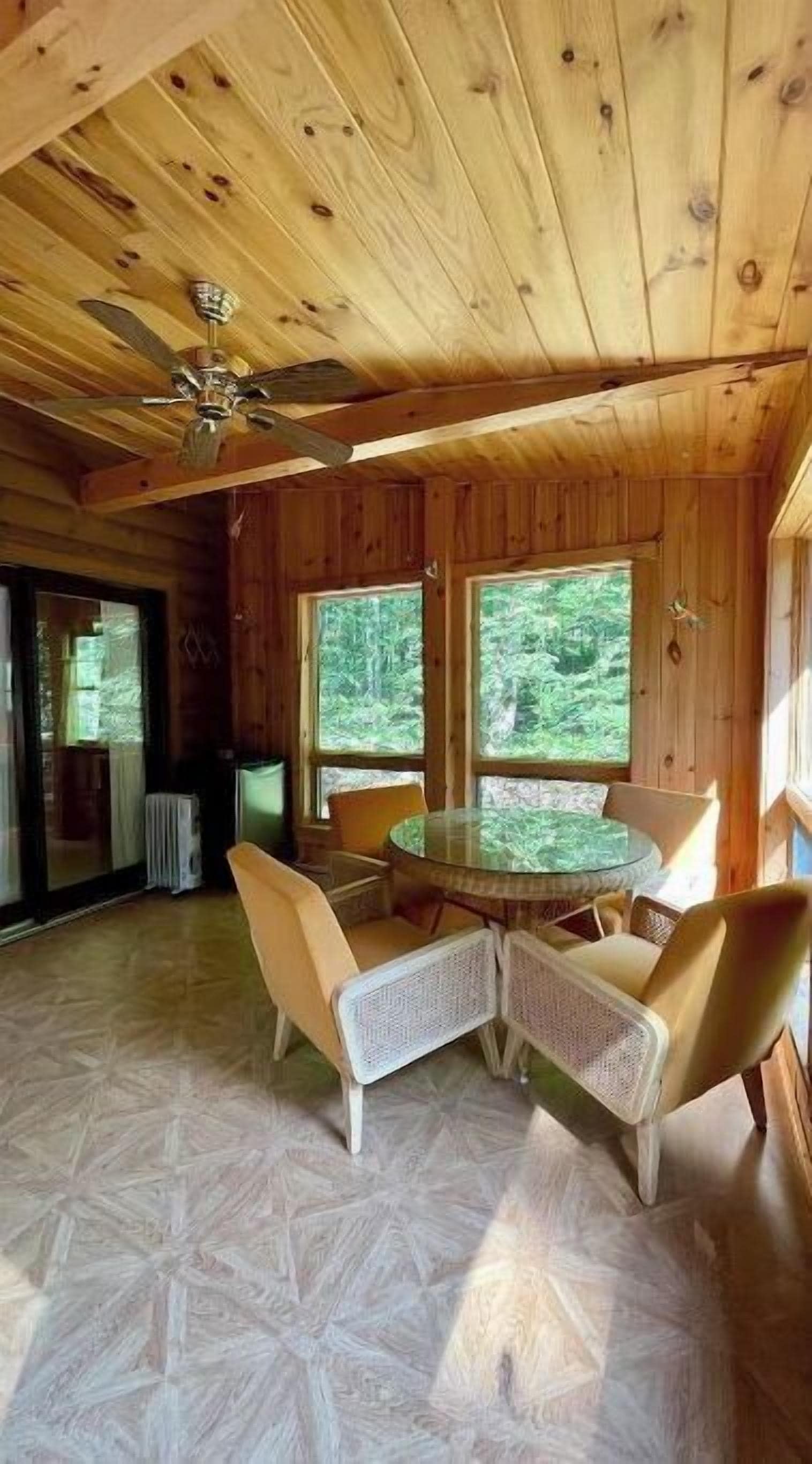 A wooden cabin interior with a round table and four chairs, large windows, and a ceiling fan.