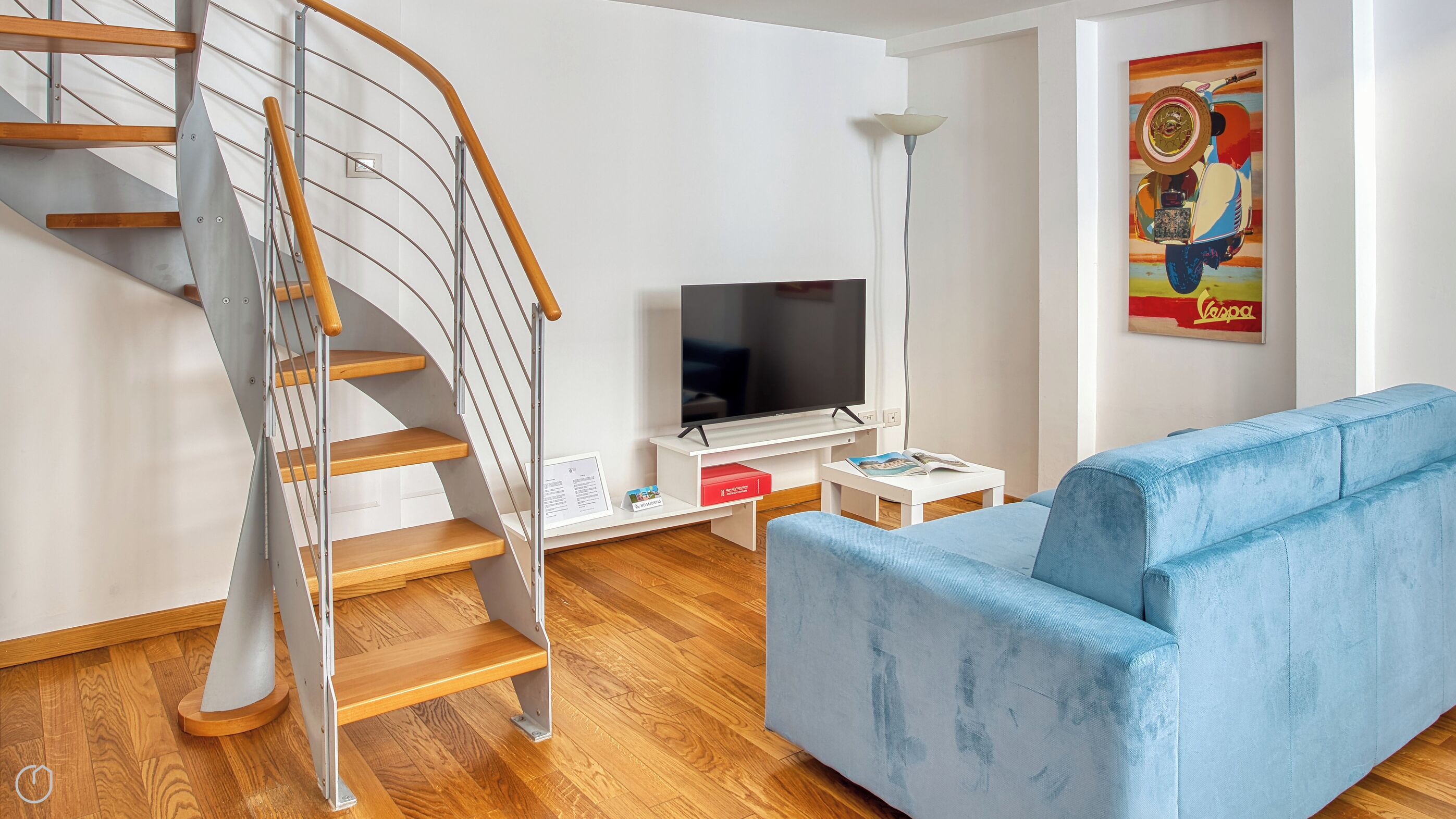 A modern living room with a wooden staircase, a flat-screen TV, a blue sofa, and a colorful artwork on the wall.