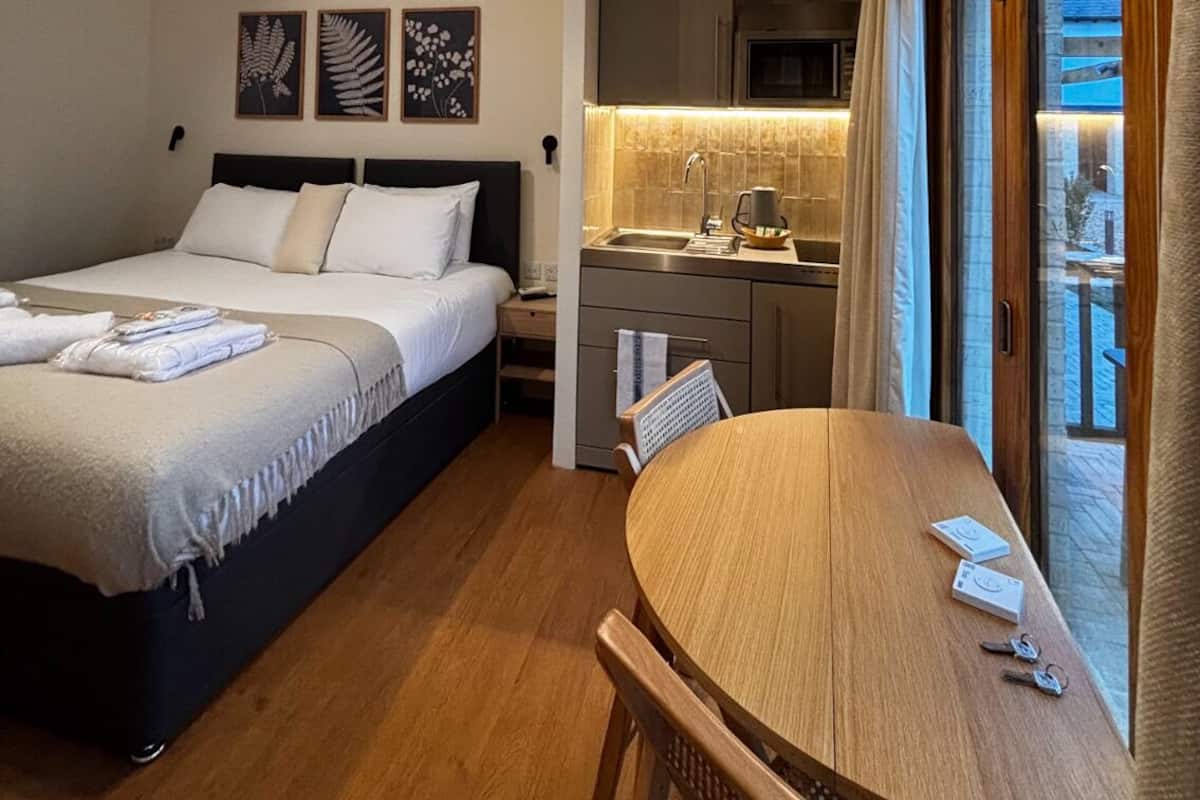 A compact hotel room with a bed, a small kitchenette, a dining table, and a view through a large window.