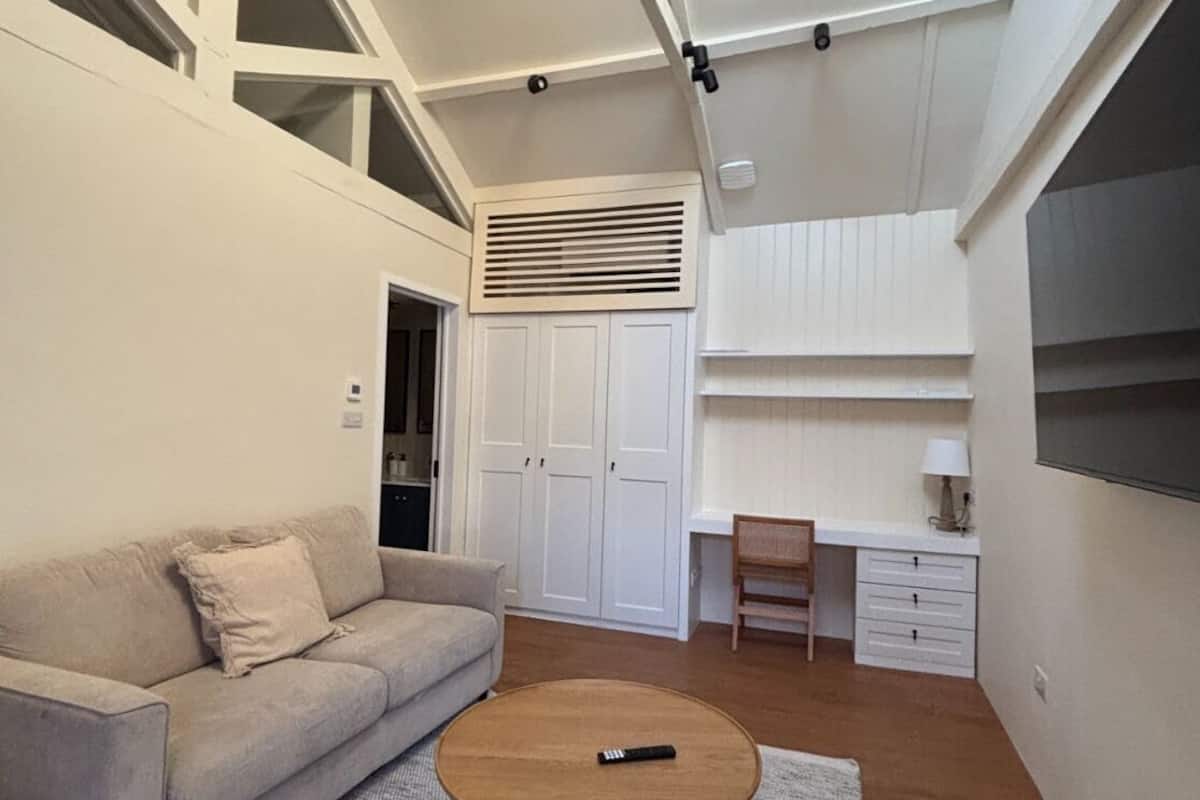 A modern living room with a sofa, a coffee table, and a desk with a chair. There is a wardrobe and a window with blinds.