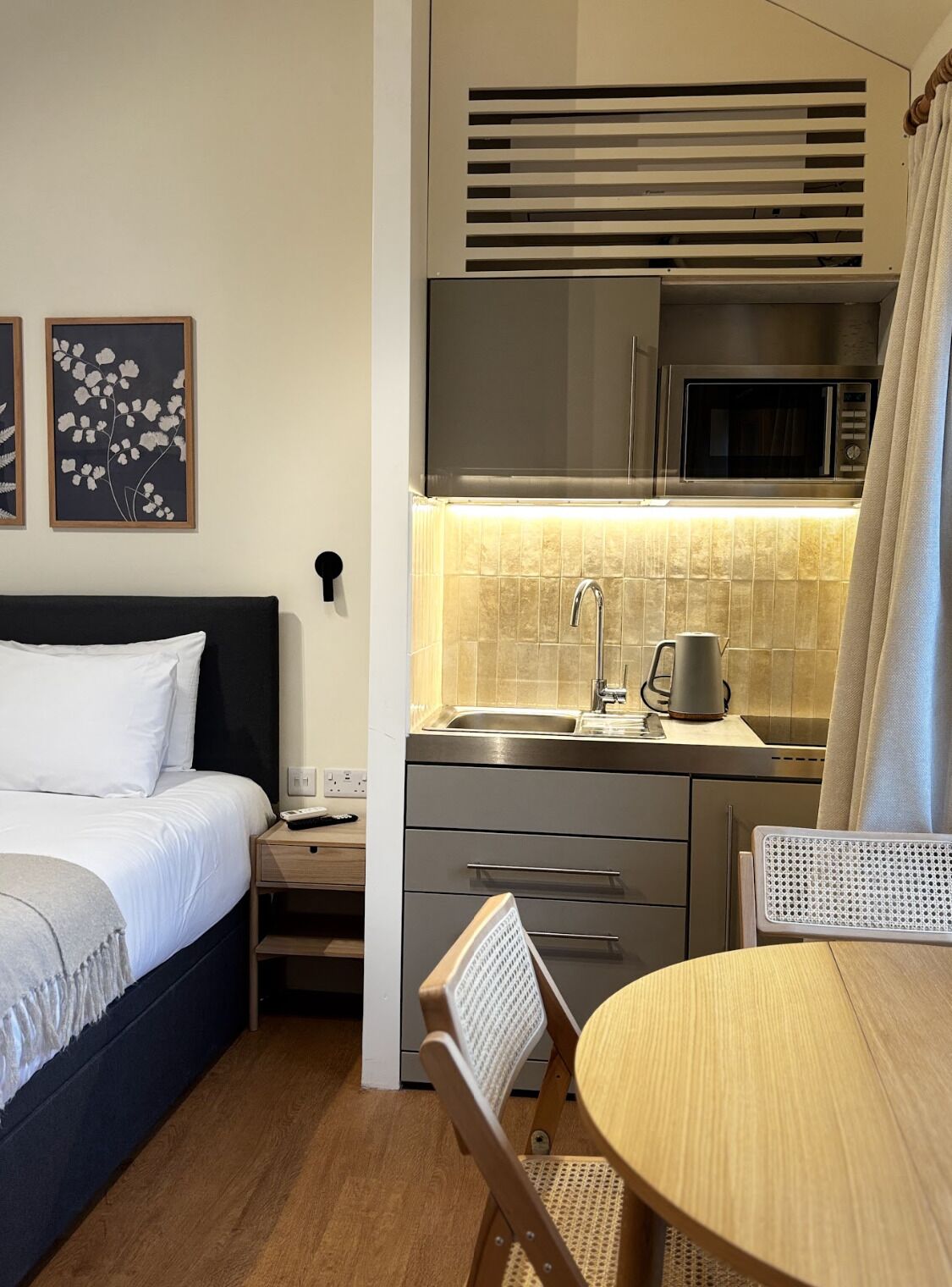 A compact hotel room with a bed, bedside table, a small kitchenette with a microwave, a sink, and a chair.