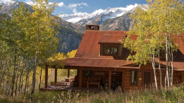 A rustic cabin with a metal roof, surrounded by trees and mountains in the background.