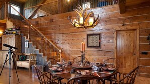 A rustic wooden interior with a dining table set for four, a chandelier, and a staircase leading to an upper level.