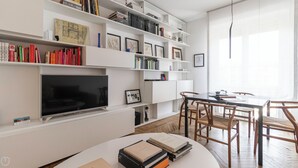 A modern living room with a flat-screen TV, white shelves with books and framed pictures, a dining area with a table and chairs, and a large window with sheer curtains. - Italianway - Palestrina 31 (Milan)