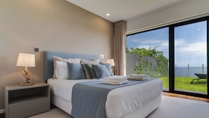 A modern bedroom with a large bed, two bedside tables, and a view of a grassy area through a large window. - Mango (Funchal)