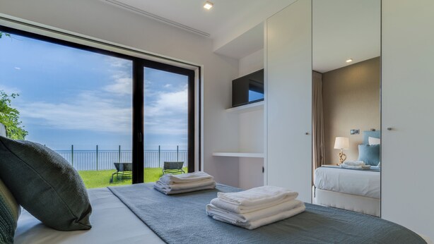 A modern hotel room with a large sliding glass door leading to a balcony, a neatly made bed, a bedside table with a lamp, and a TV mounted on the wall. - Mango (Funchal)