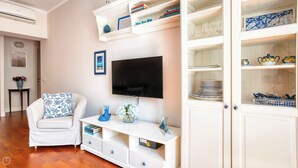 Un salon avec un canapé blanc, un téléviseur à écran plat, une console blanche agrémentée d’objets décoratifs et un meuble vitré contenant de l’ustensile de cuisine.