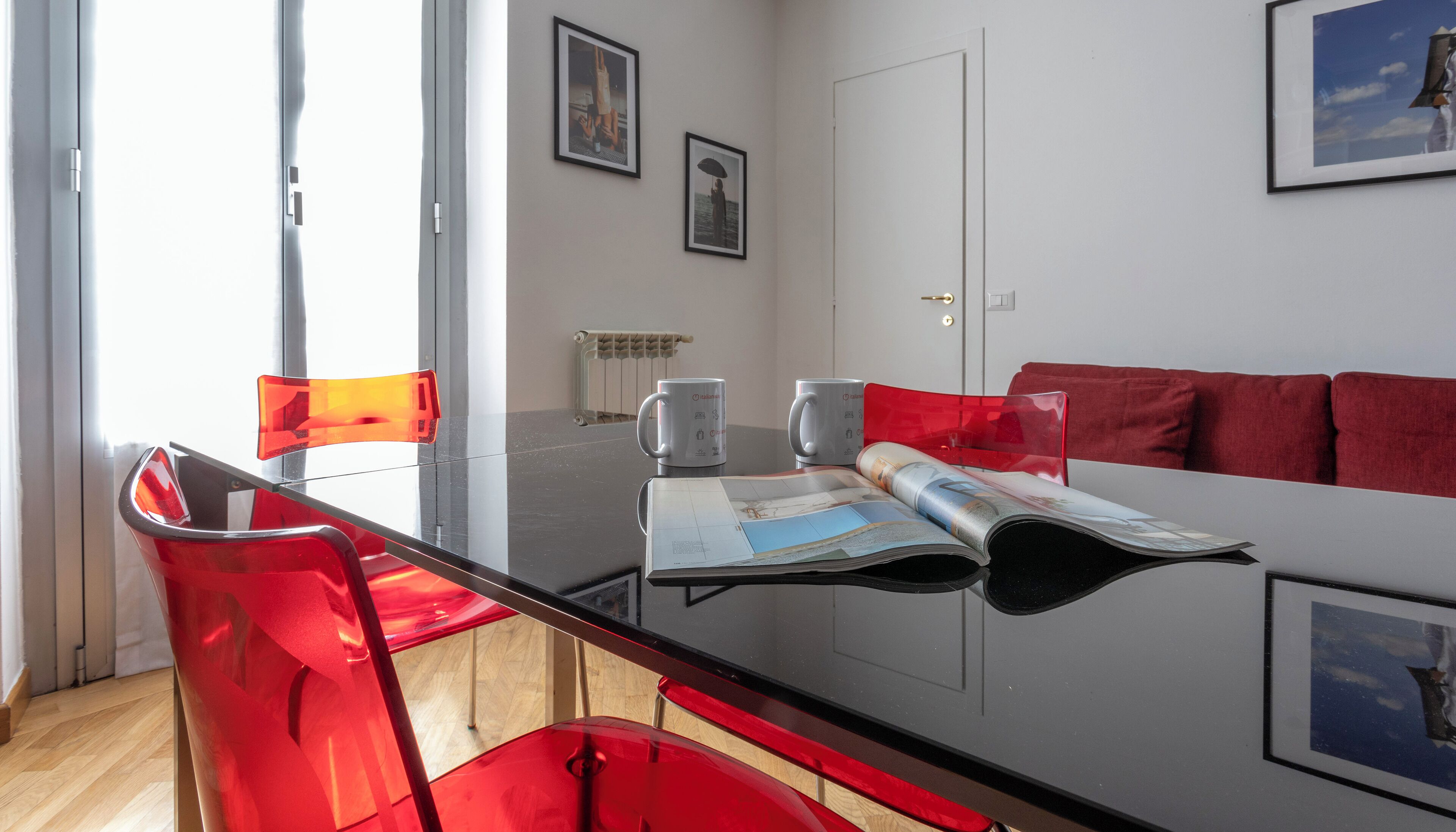 Uma sala de estar moderna com uma mesa de jantar de vidro, cadeiras vermelhas, um sofá vermelho e duas fotos emolduradas na parede.