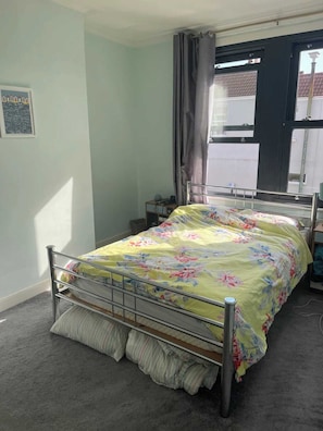 Une chambre à coucher avec un lit en métal, une couvre-lit à motifs floraux, une table de chevet avec une lampe et une fenêtre avec des rideaux.