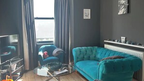 Ein Wohnzimmer mit einem petrolfarbenen Sofa, einem Glastisch, einem Fernseher und einer Heizung.