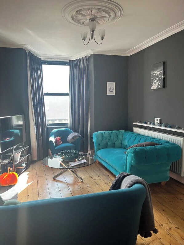 A living room with a teal sofa, a glass coffee table, a TV, and a radiator.