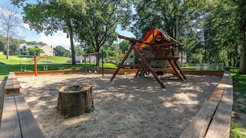 Un parque infantil con columpios y una estructura de escalada, arena, árboles y una mesa de picnic.