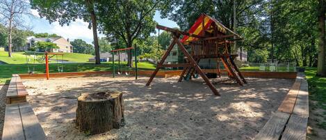 En legeplads med gyngestativ og klatrestruktur i en park med sand, træer og et picnicbord.